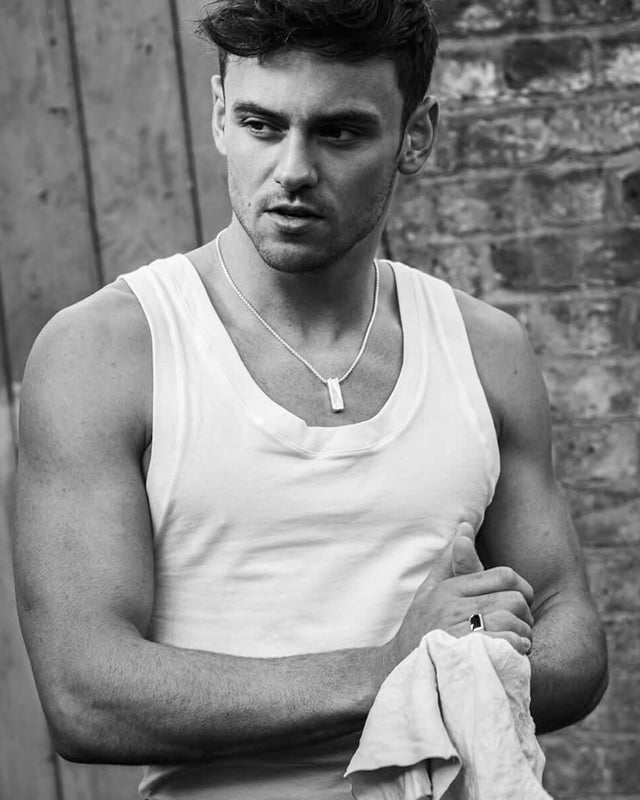 Tom Daley wearing a white tank top, a silver necklace and an inlaid oxford ring with onyx against a brick wall